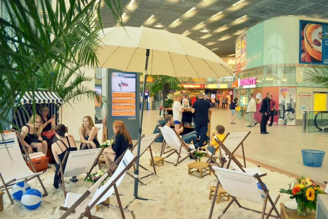 Reisende entspannen in Liegestühlen auf der Sandfläche in der Bahnhofshalle